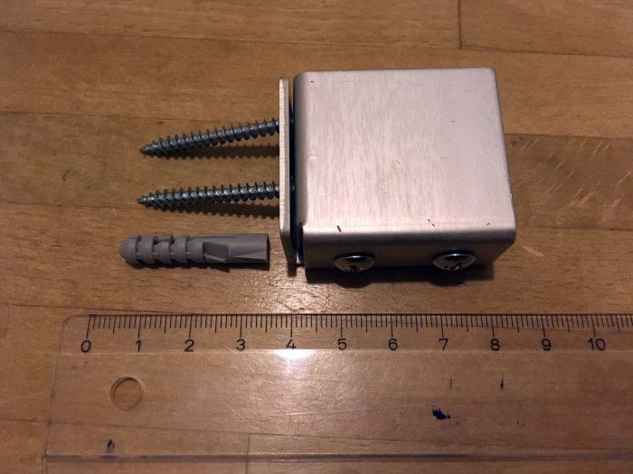 Metallfäste för duschdraperiskena med två skruvar och en plugg bredvid en linjal på ett träbord.