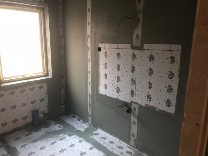 Ett ofärdigt rum med isoleringsplattor på väggarna, synliga rör och ledningar, vid ett husbygge.