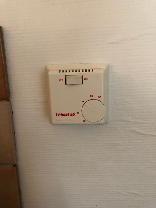 En väggmonterad, analog termostat för värmereglering märkt e-I heat ab, med temperaturvred och on/off-switch.