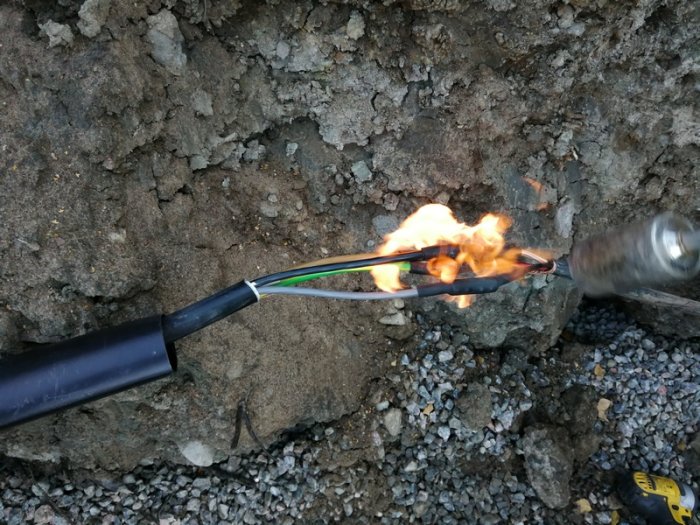 Elektrisk kabel som svetsas med en gasbrännare utomhus med jord och grus i bakgrunden.