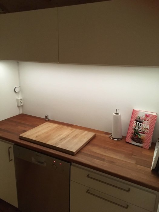 Kök med nya skåp och arbetsbänk, med belysning under överskåpen och en diskmaskin samt en träskärbräda och köksbok på bänken.