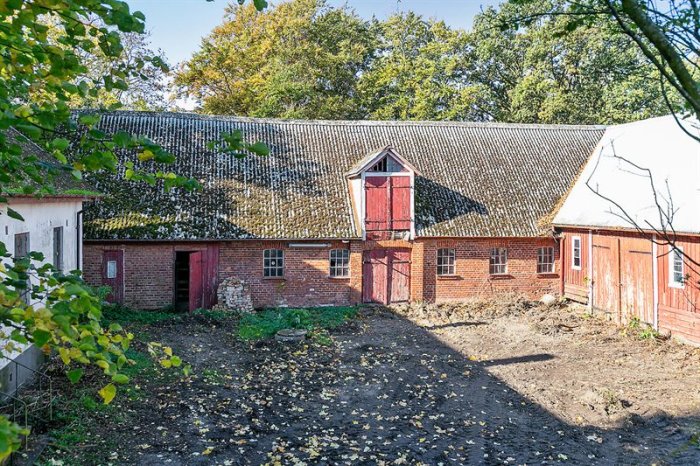 Stort gammalt bostadshus i tegel med slitna röda dörrar och måsstensbelagd tak, omgivet av träd och löv.