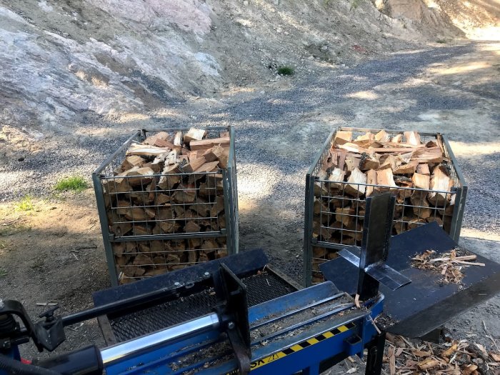 Två fyllda vedstaplar i metallkorgar och en blå vedklyv framför en grusig bakgrund.