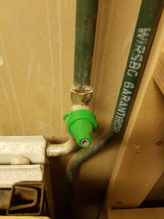 Grön plastkåpa märkt 'Danfoss' på en ventil till en vattenburen radiator, fastsatt på rör.
