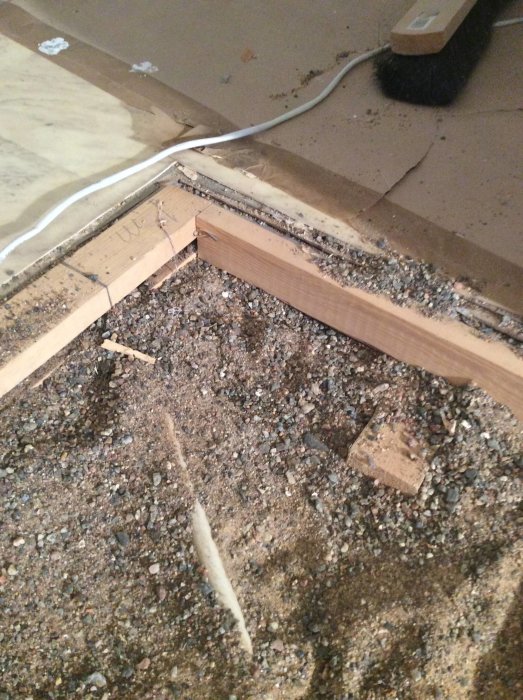 Golvsituation med synliga reglar, elkabel i vitt rör, kilar och grusigt underlag under pågående renovering.