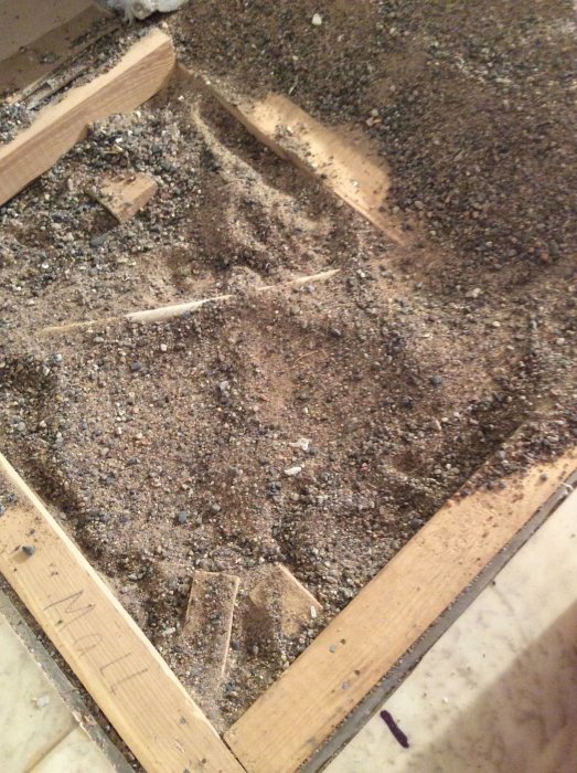 Avtäckt golv visar grov grus och sand, med träreglar, kilar och synligt vitt elkabelskyddsrör.