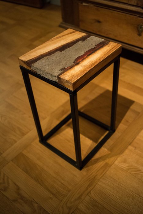 Handgjord sidobord med en topp av trä och inlägg av betong, på ett svart metallunderrede, stående på ett parkettgolv.