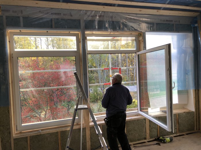 Person i byggarbetskläder står på stege och installerar fönster i ett halvfärdigt rum med isolerade väggar och synlig träkonstruktion.