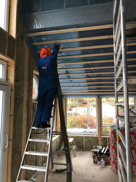 Person i byggkläder gipsar taket i ett byggprojekt med isoleringsmaterial på väggarna och byggmaterial på golvet.