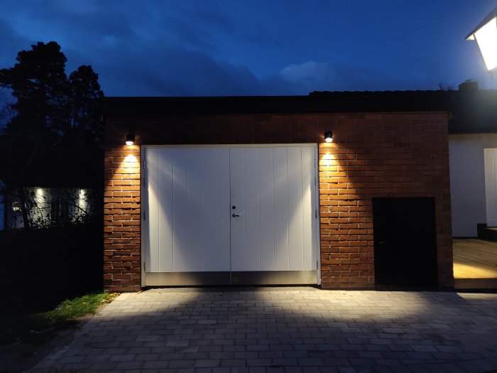 Belysningsarmaturer monterade ovanför en vit garageport i tegelhus under skymningen.