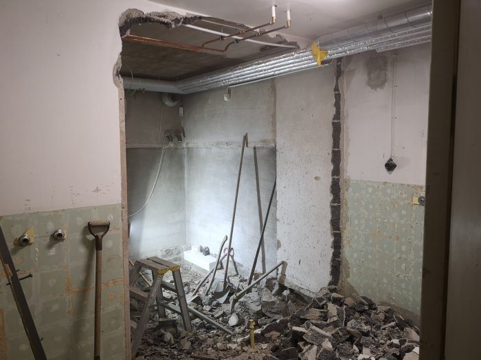 Rivningsarbete inne i ett rum med synliga bergstycken, rördragning i taket, och en kanalfläkt till höger.