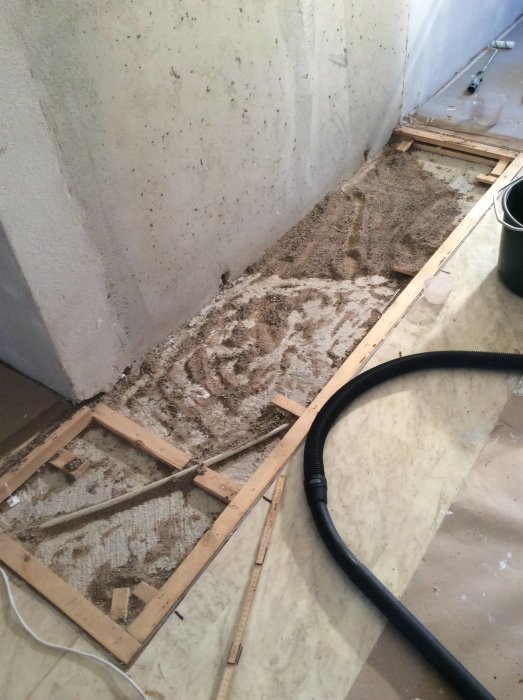 Renoveringsprojekt med uppbrutna träreglar och sand på ett ojämnt betonggolv nära en vägg.