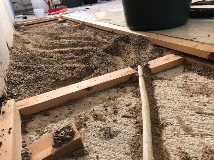 Öppnat golv med synligt sandlager och träreglar mot en ojämn betongvägg under en renoveringsprocess.