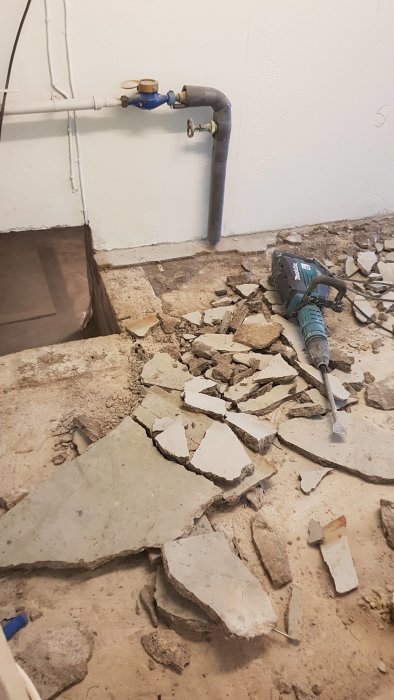 Upptaget betonggolv med trasiga plattor och en borrhammare nära vattenrör i en tvättstuga.
