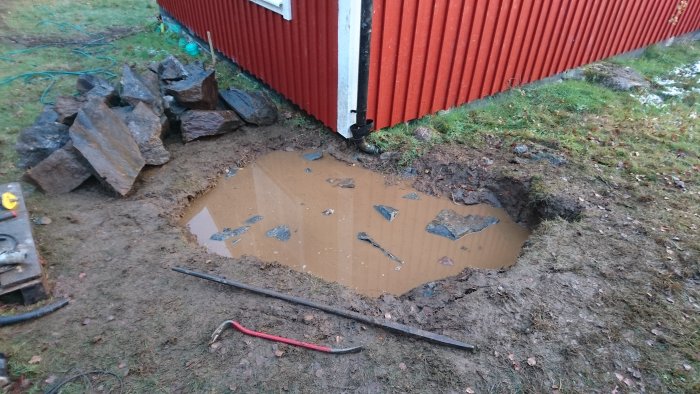 Grävd grop vid husvägg full av vatten, med stenar och verktyg runtomkring.