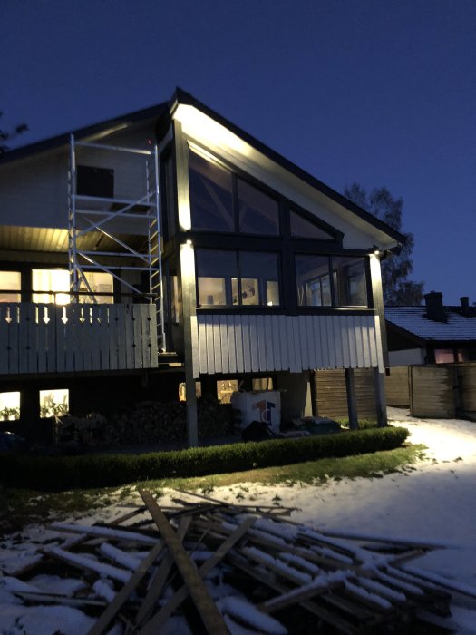 Ett hus i skymningen med en nyinstallerad utelampa som är tänd, snö på marken och byggställning vid fasaden.