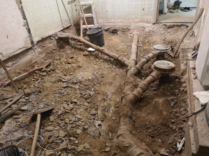 Rivet betonggolv som exponerar gamla avloppsrör och rörledningar i en byggarbetsplats inomhus.