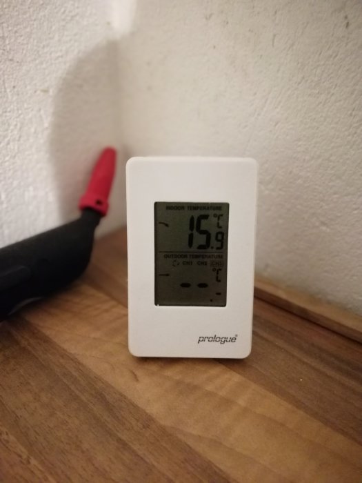 Termometer visar inomhustemperatur på 15,8 grader ståendes på ett trägolv med en verktygsspets i bakgrunden.