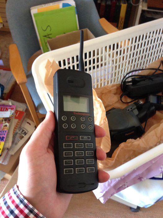 En hand håller en vintage Technophone mobiltelefon med utdragen antenn och stort batteri, bakgrund av rörigt rum.