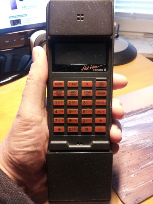 Hand som håller en vintage Ericsson Hotline-mobiltelefon från sent 80-tal framför en dator och tangentbord.