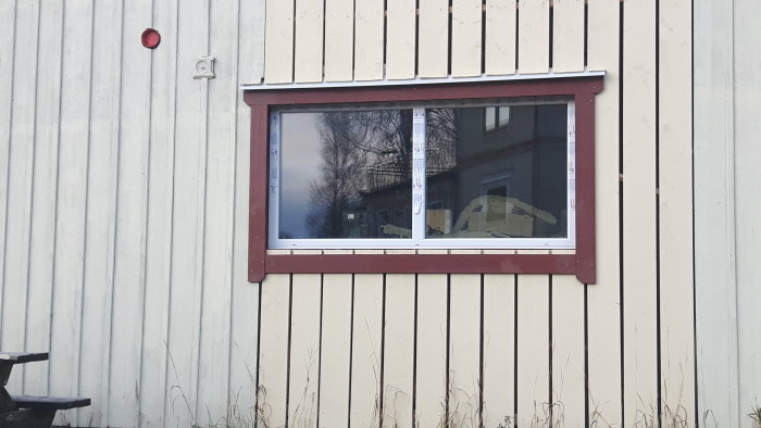 Ett nyinstallerat fönster med rödbrun omramning på en ljusbeige plåtfasad, reflektioner syns i glaset.