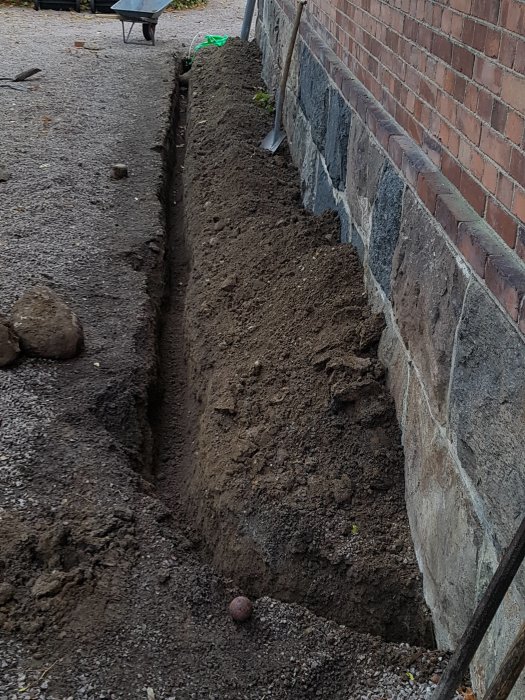 Grävd grävning längs en husvägg för fiberinstallation med spade och skottkärra i bakgrunden.