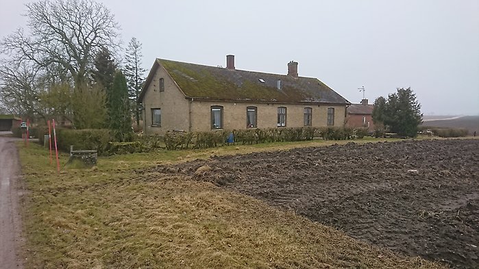 Långt gammalt hus med mossigt tegeltak, omgivet av en nyplogad åker och kala träd under en mulet himmel.