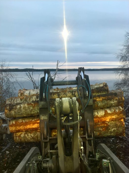 Gripklo lyfter stockar av björkämnen med en sjö och skog i bakgrunden under en klar himmel.