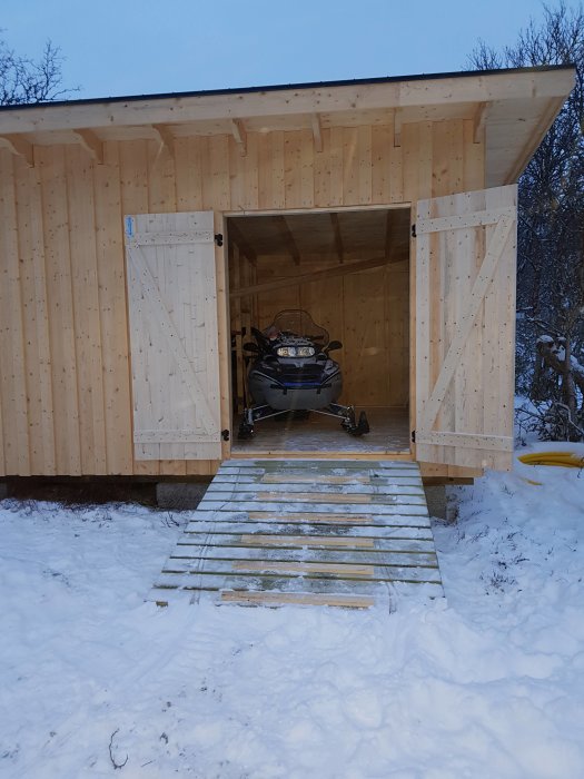 En snöskoter parkerad i ett öppet träskjul med en trärampramp i snöigt landskap.
