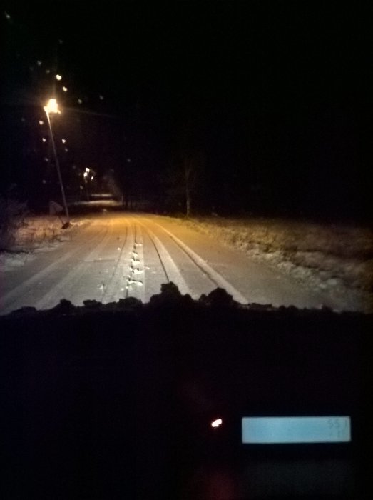 Utsikt från ett fordon vid gryningen med snöflingor som faller och upplyst snötäckt väg framför.