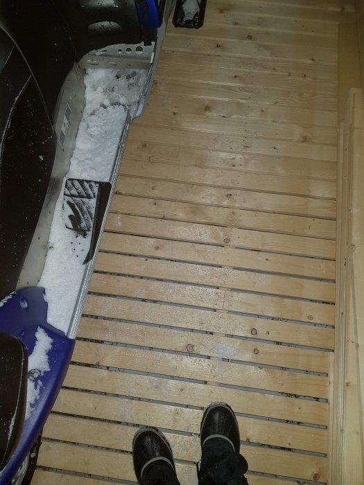 Person står på ett nybyggt trägolv i en carport med en bil delvis täckt av snö till vänster.