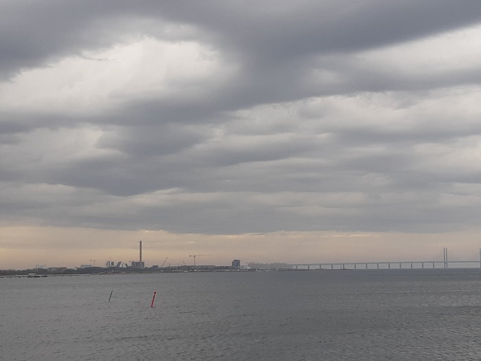 Molnig himmel över en bro och industribyggnader i fjärran med spegling i vattnet.