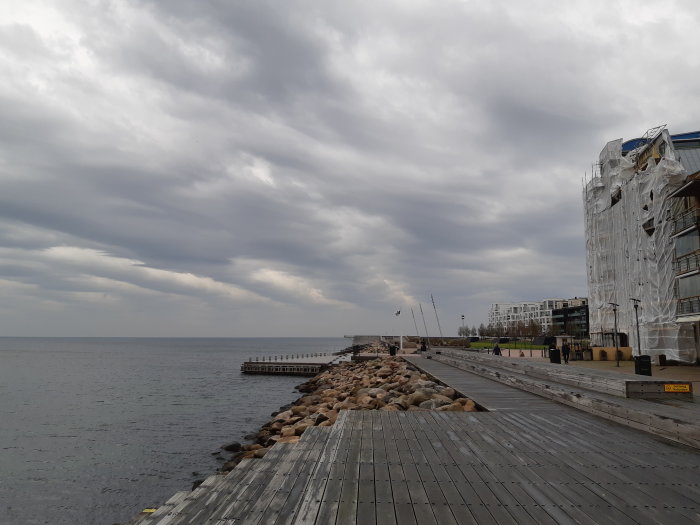 Molnig himmel över en promenadbrygga vid vattnet med stenar och en byggnad inpackad i skyddsplast.