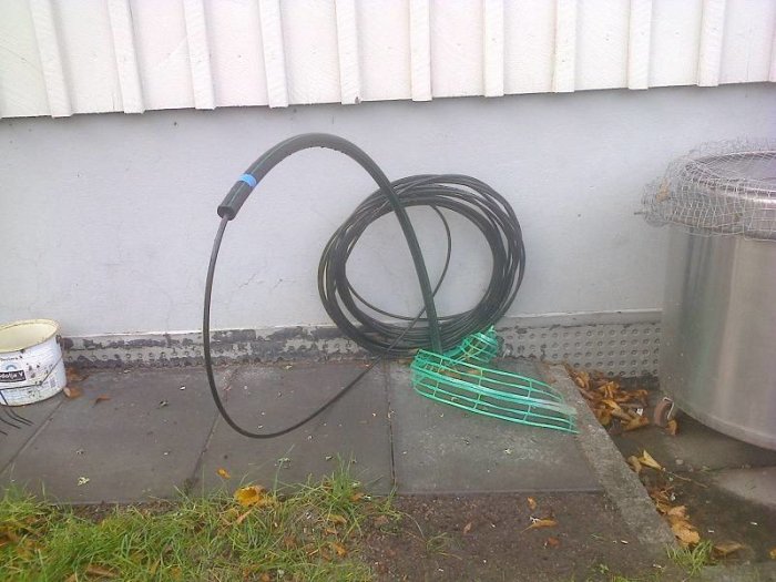 En rulle med svart kabel upplagd mot en husvägg bredvid en vattentunna och fallna löv.