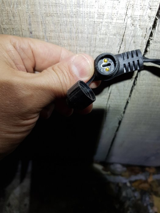 Hand håller en elektrisk kontakt med synliga stift framför trävägg.