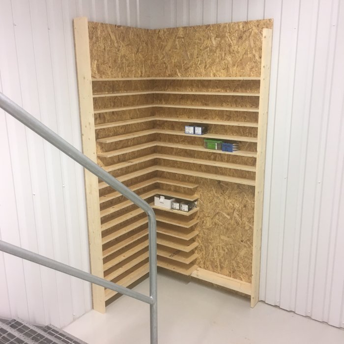 Hörnhylla med flera grunda hyllplan fylld med få skruvförpackningar i ett ljust rum med vit vägg och träpanel.