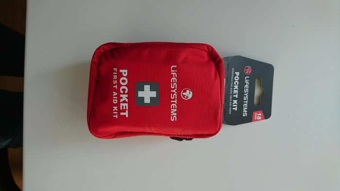 Röd första hjälpen-kit, märkt "LIFESYSTEMS POCKET FIRST AID KIT", ligger på ett vitt bord.