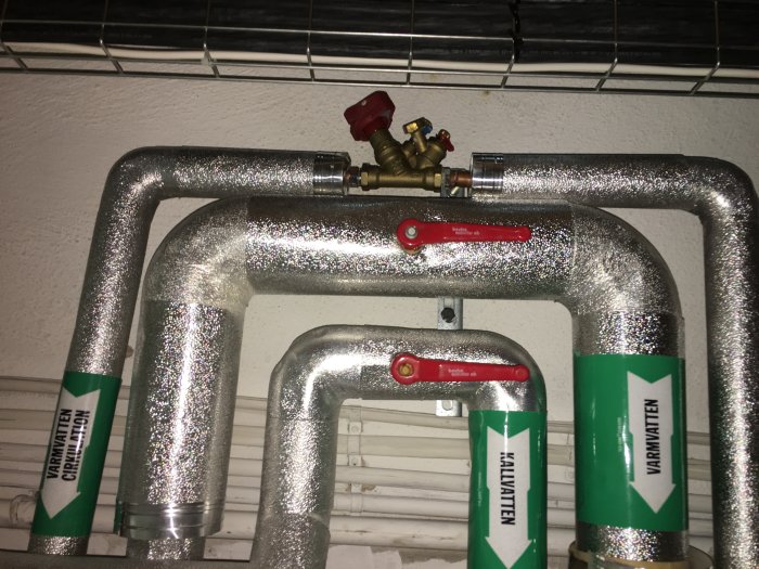 Isolerade rör med röda ventiler och etiketter för varmvatten och cirkulation.