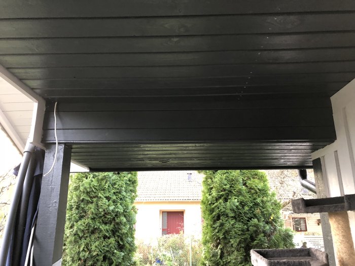 Svartmålad träpanel under takutskjut med synligt rör och stolpar, utsikt mot trädgård.