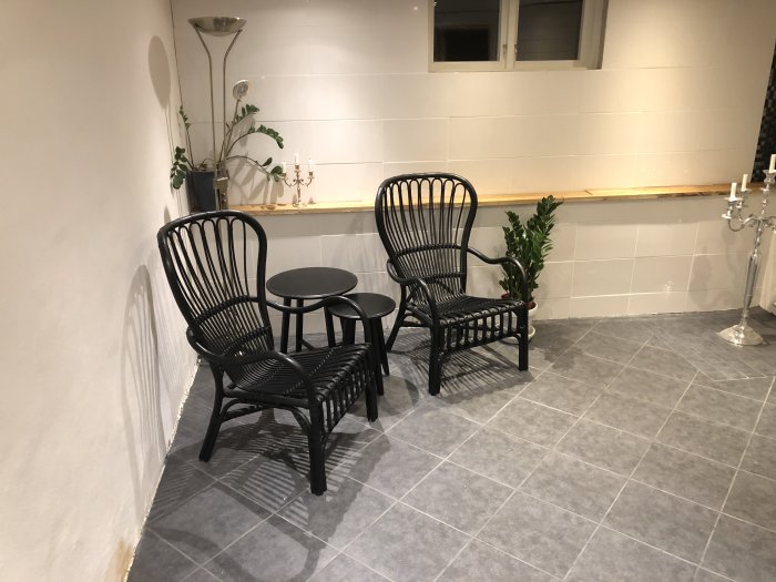 Nyinredd relaxdel med två svarta stolar och ett runt bord på gråa klinkers, vit vägg med hylla och växter.