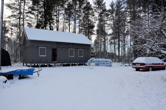 Hus under konstruktion med isoleringsskivor och läkt på gaveln, omgivet av snötäckt terräng, skog och bilar med snö på.