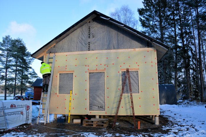 En person som sätter upp isoleringsskivor på en husgavel, med snö och byggmaterial runtomkring.