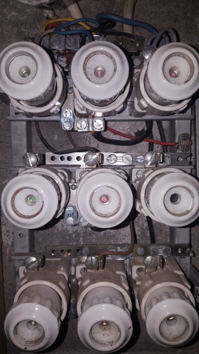 Gammal elektrisk säkringstavla med keramiska säkringshållare och anslutna kablar i ett hus.