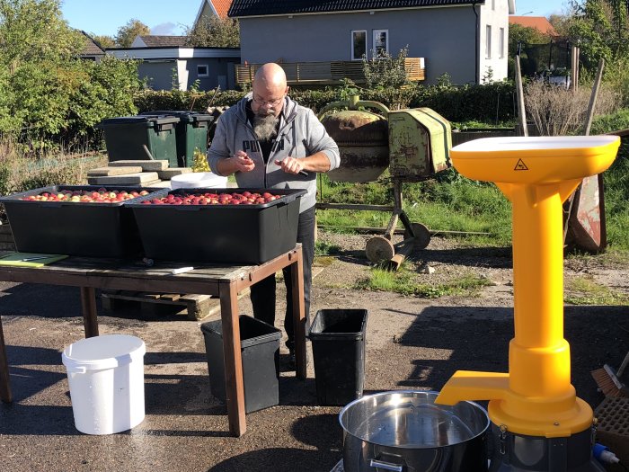 Person rensar äpplen vid ett utomhusbord nära äppelmustpress på en solig dag.