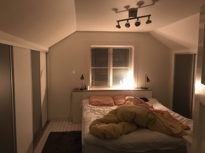 Sovrum med snedtak, inbyggda garderober, upplyst fönster med persienner och belyst säng med gult täcke.