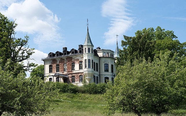 Förfallen historisk byggnad med torn som liknar ett slott, omgiven av gröna träd och buskar, under renovering.