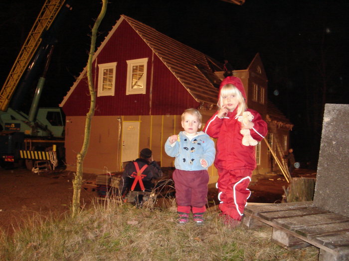 Två små barn framför ett rött hus som lyfts av en kran under kvällstid.