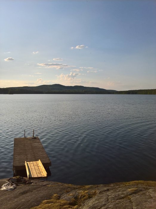 En brygga som sträcker sig ut i lugnt vatten med skogklädda kullar i bakgrunden mot en klar himmel.