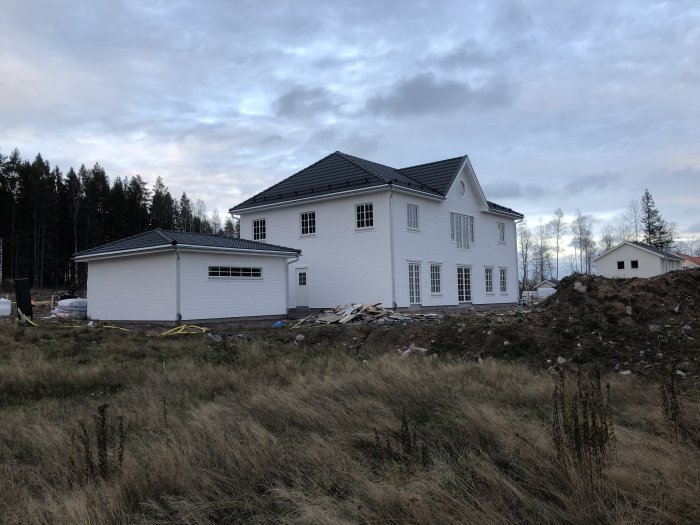 Nybyggt vitt hus med svart tak och angränsande garage i ett landskap under konstruktion.