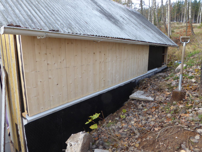 Nyinstallerad vindskyddspapp och panel på husvägg med plåt vid grundmuren och spade i förgrunden.